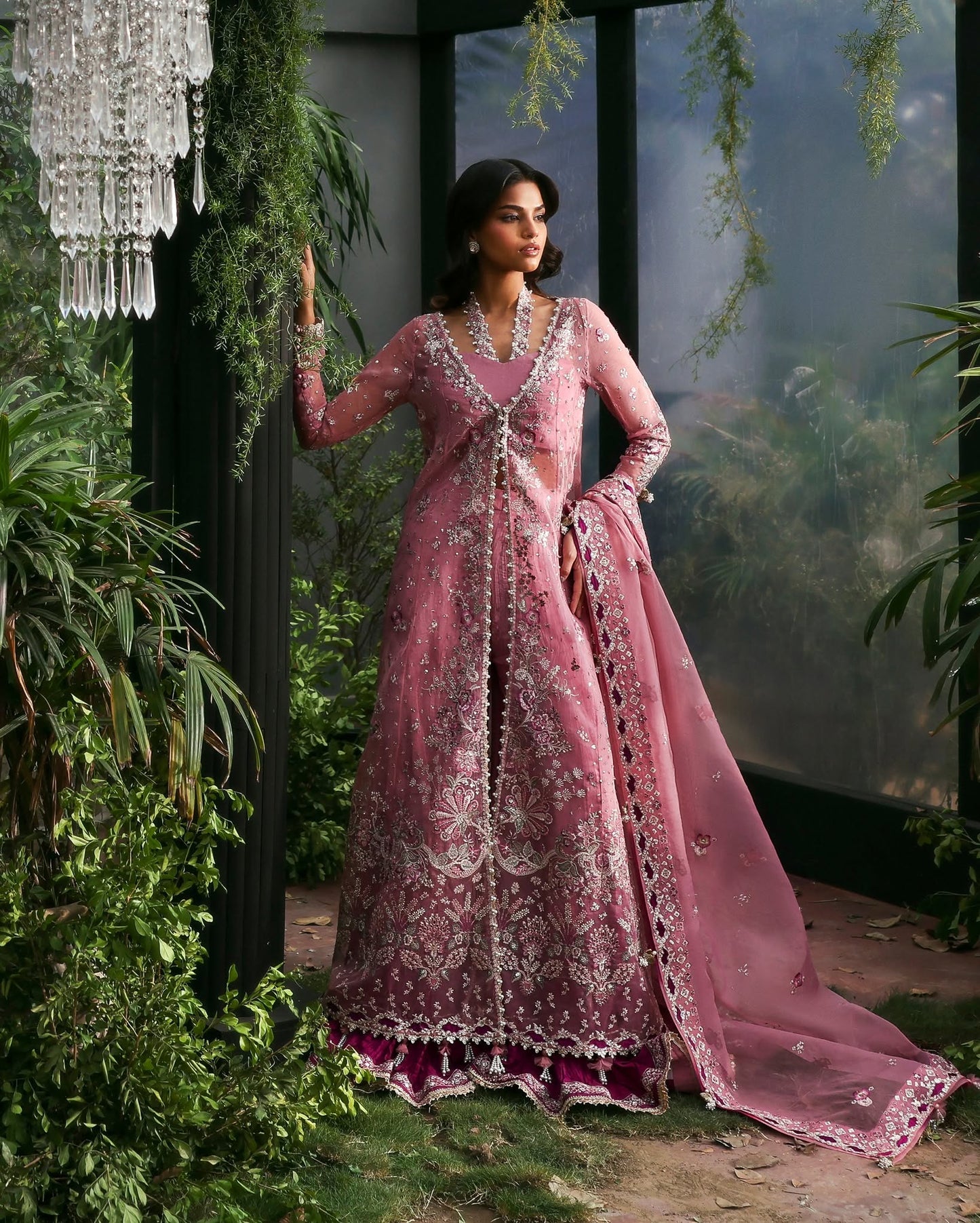 Woman in a pink embroidered traditional outfit standing in a decorative setting with plants and a chandelier.