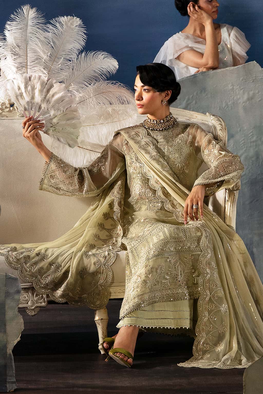 Woman in an ornate dress with a feathered fan, sitting on a chair against a blue background.