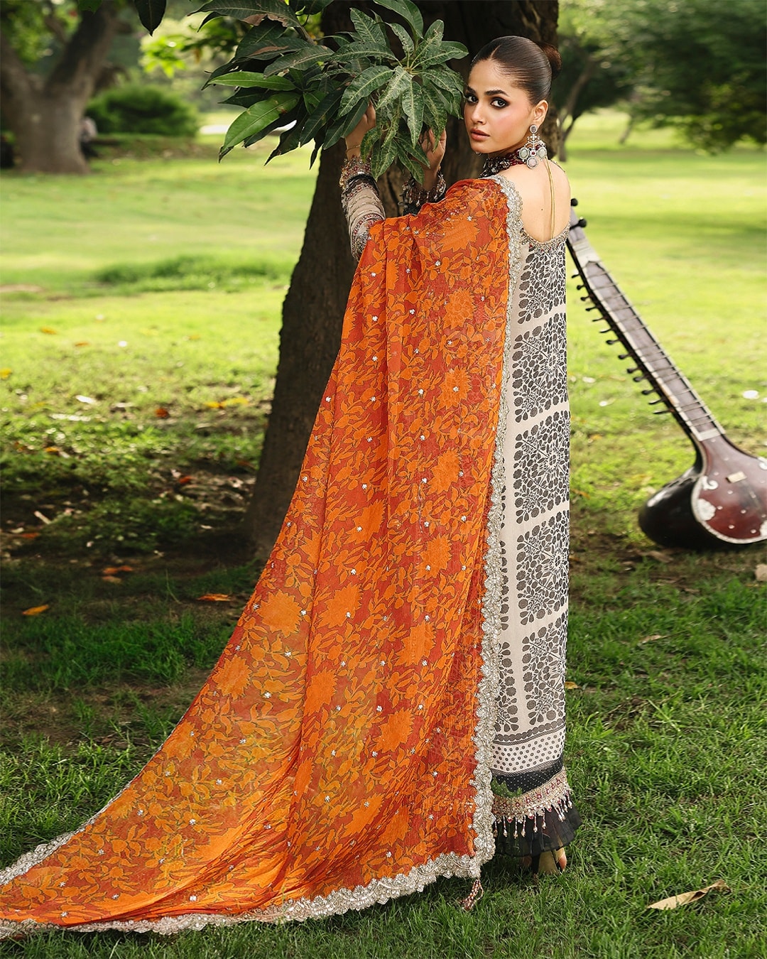 Woman in an orange saree with a white border standing outdoors on grass.