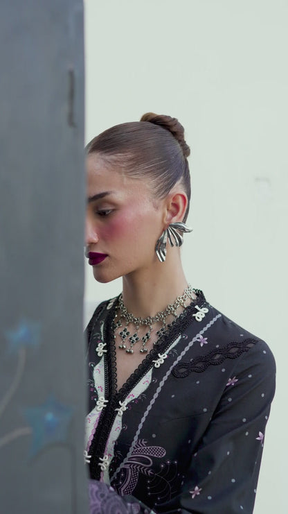 Woman wearing a black outfit with intricate designs and butterfly earrings against a neutral background