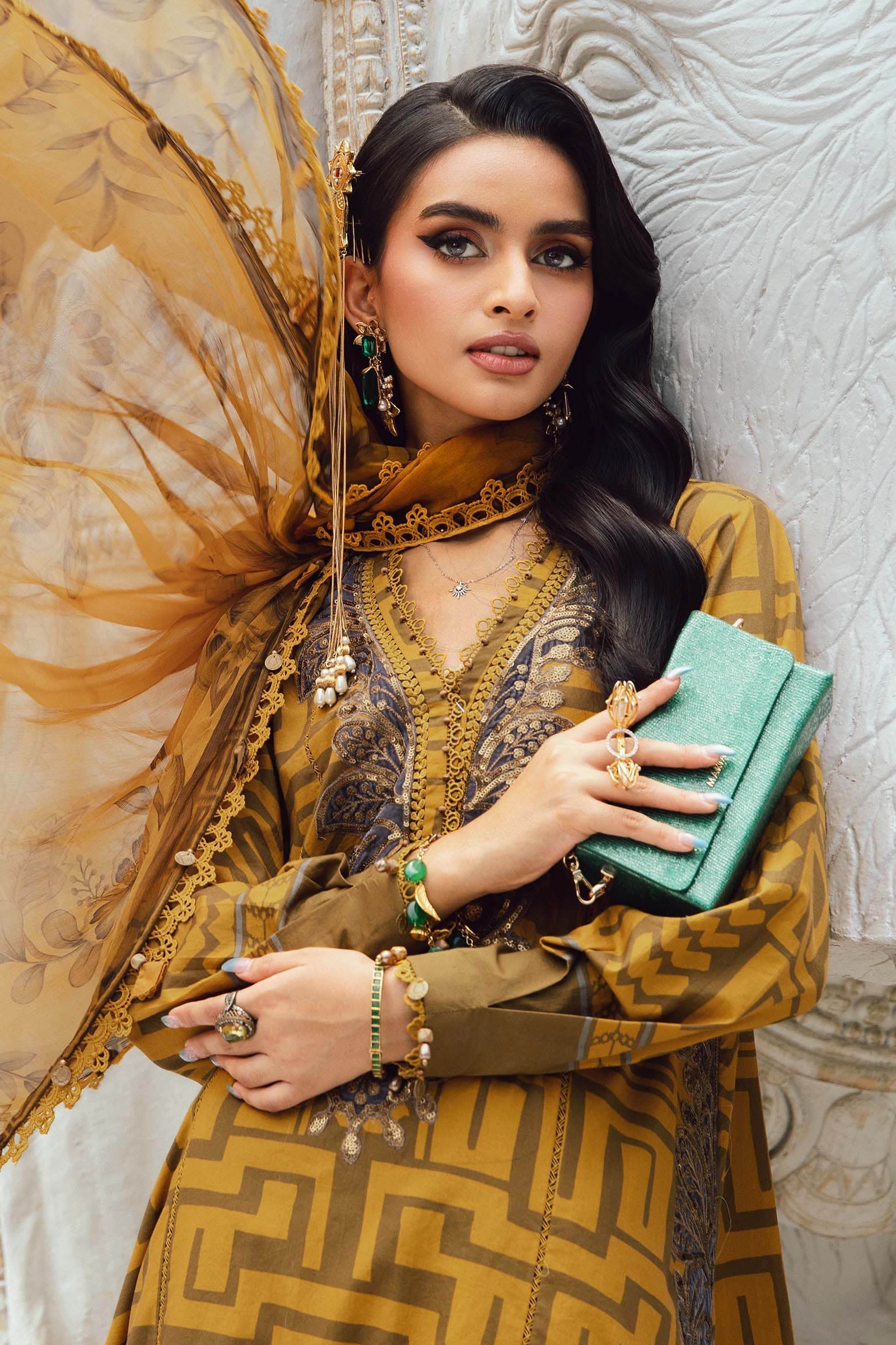 Woman in traditional outfit holding a green clutch against a textured wall.